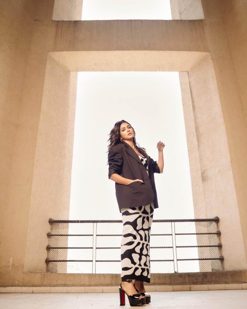 A woman in a black blazer and patterned pants standing in front of a building.