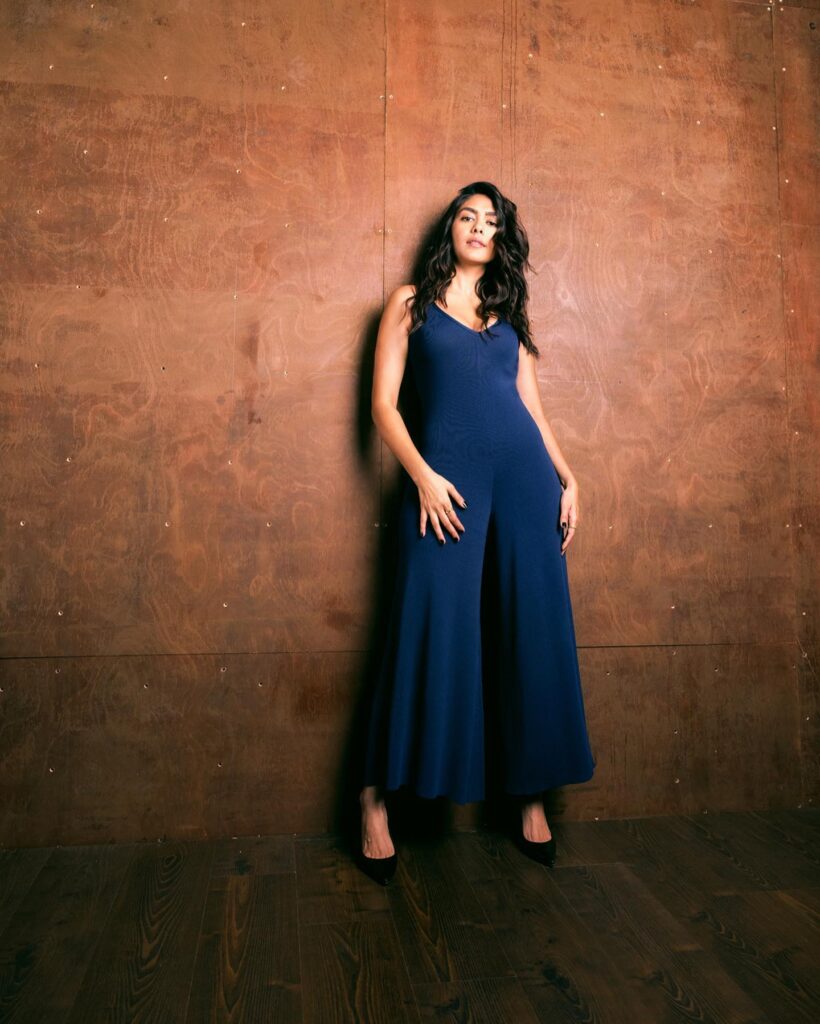 A woman in a blue jumpsuit posing against a brown wall.